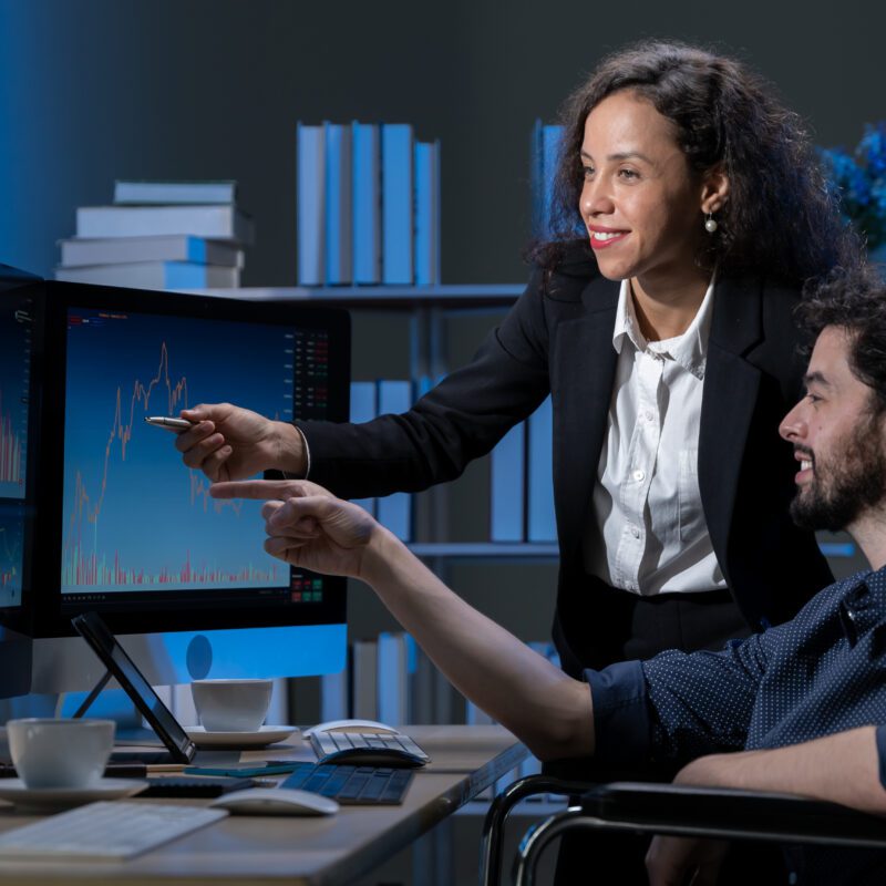 Two professionals analyze financial charts on computer screens in a modern office. One points at the screen, both displaying engaged teamwork.
